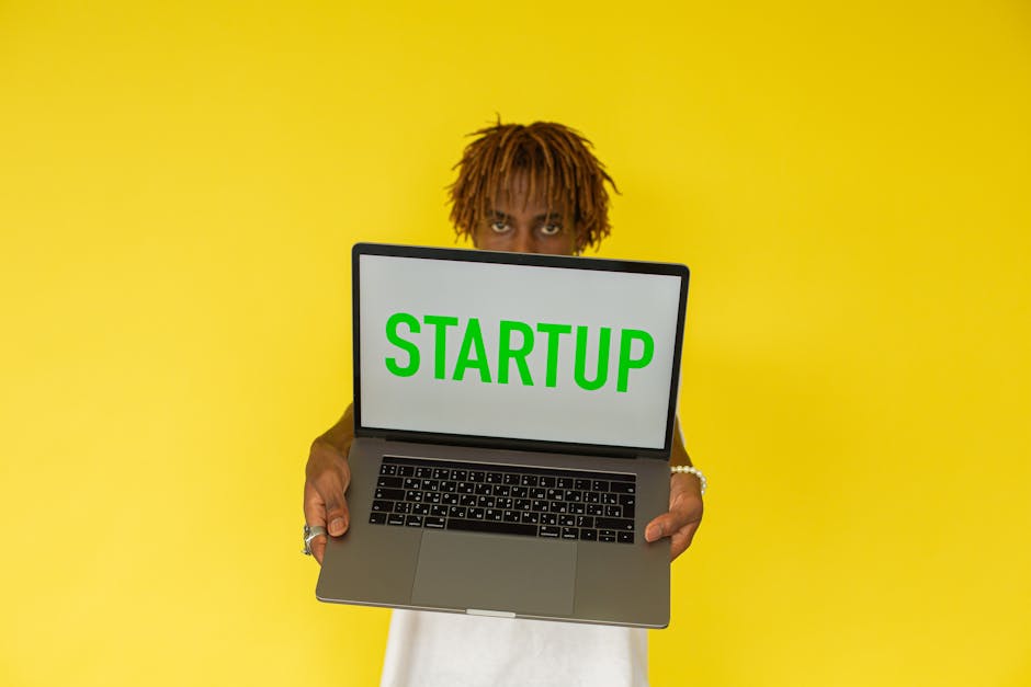 A young black man holds a laptop displaying 'Startup' against a vibrant yellow background.